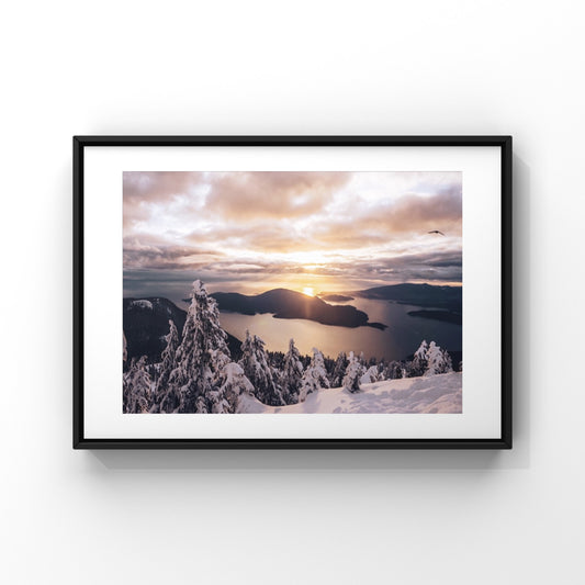 Mountain sunset photography print of the north shore mountains in Vancouver, British Columbia. The sky is peach, yellow and pink, the trees are covered in snow, there is a bird flying in the sky.